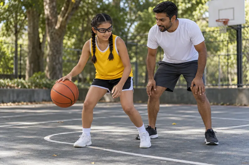 A child wearing protective sports goggles practices basketball with an adult coach outdoors.